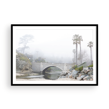Landscape coastal photography of a concrete arch bridge disappearing into a dense fog, flanked by palm trees and rocky shores. This calm beach print evokes a mysterious seascape. Photograph in minimalist mock up.
