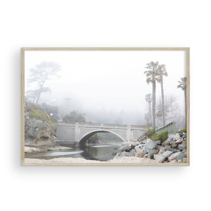 Landscape coastal photography of a concrete arch bridge disappearing into a dense fog, flanked by palm trees and rocky shores. This calm beach print evokes a mysterious seascape. Photograph in minimalist mock up.