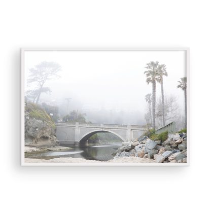 Landscape coastal photography of a concrete arch bridge disappearing into a dense fog, flanked by palm trees and rocky shores. This calm beach print evokes a mysterious seascape. Photograph in minimalist mock up.