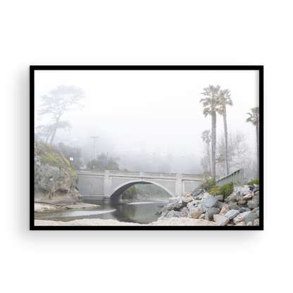 Landscape coastal photography of a concrete arch bridge disappearing into a dense fog, flanked by palm trees and rocky shores. This calm beach print evokes a mysterious seascape. Photograph in minimalist mock up.