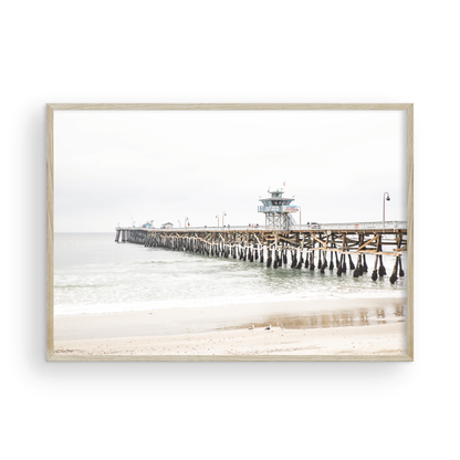 A long wooden pier, the San Clemente Pier, extends into the ocean in this landscape coastal photography. The beach in the foreground suggests a calm ocean print, ideal for a large coastal wall art or a beach cottage wall decor. Photograph in minimalist mock up.