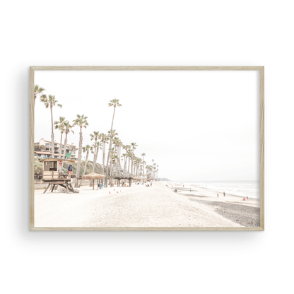 A wide landscape coastal photography view of San Clemente State Beach in Southern California, featuring a row of tall palm trees, a lifeguard tower, and people enjoying the calm beach. This horizontal image evokes Summer Vibes Wall Art and tropical beach print aesthetics. Photograph in minimalist mock up.
