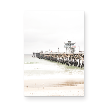 A long wooden pier, the San Clemente Pier, extends into the ocean in this vertical coastal photography. The beach in the foreground suggests a calm ocean print, ideal for a large coastal wall art or a beach cottage wall decor. Photograph in minimalist mock up.