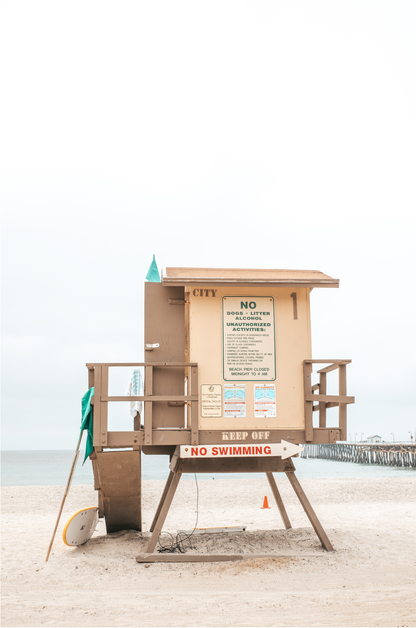 A vertical beach art close-up of a rustic lifeguard tower on San Clemente Beach, with a surfboard leaning against it and the pier visible in the distance. This image is perfect for a minimalist surf wall decor, surf nursery decor, or a fun surf print.