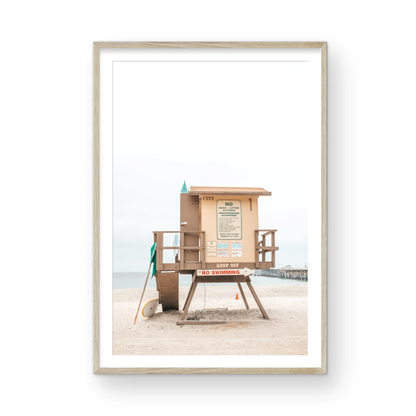 A vertical beach art close-up of a rustic lifeguard tower on San Clemente Beach, with a surfboard leaning against it and the pier visible in the distance. This image is perfect for a minimalist surf wall decor, surf nursery decor, or a fun surf print. Photograph in minimalist mock up.