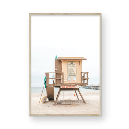 A vertical beach art close-up of a rustic lifeguard tower on San Clemente Beach, with a surfboard leaning against it and the pier visible in the distance. This image is perfect for a minimalist surf wall decor, surf nursery decor, or a fun surf print. Photograph in minimalist mock up.