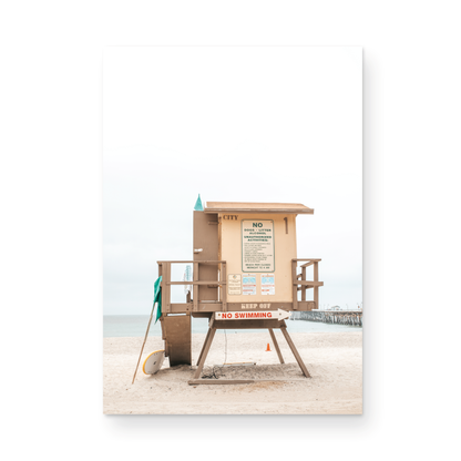 A vertical beach art close-up of a rustic lifeguard tower on San Clemente Beach, with a surfboard leaning against it and the pier visible in the distance. This image is perfect for a minimalist surf wall decor, surf nursery decor, or a fun surf print. Photograph in minimalist mock up.