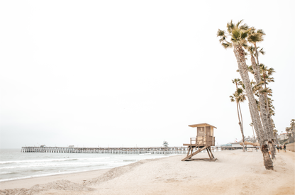 A wide landscape coastal photography view of San Clemente State Beach in Southern California, featuring a row of tall palm trees, a lifeguard tower, and people enjoying the calm beach. This image evokes Summer Vibes Wall Art and tropical beach print aesthetics. 