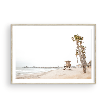 A wide landscape coastal photography view of San Clemente State Beach in Southern California, featuring a row of tall palm trees, a lifeguard tower, and people enjoying the calm beach. This image evokes Summer Vibes Wall Art and tropical beach print aesthetics. Photograph in minimalist mock up.