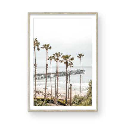 A vertical landscape coastal photography shot of the San Clemente Pier from behind a lush line of tall palm trees. This beachscape print highlights the California coast and is ideal for large beach art or a beach theme wall decor, with subtle ocean waves. Photograph in minimalist mock up.