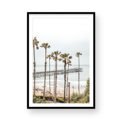 A vertical landscape coastal photography shot of the San Clemente Pier from behind a lush line of tall palm trees. This beachscape print highlights the California coast and is ideal for large beach art or a beach theme wall decor, with subtle ocean waves. Photograph in minimalist mock up.