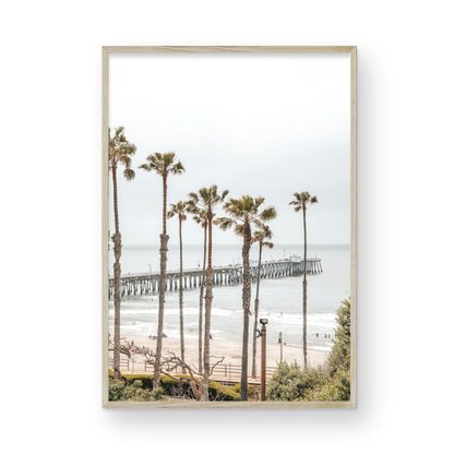 A vertical landscape coastal photography shot of the San Clemente Pier from behind a lush line of tall palm trees. This beachscape print highlights the California coast and is ideal for large beach art or a beach theme wall decor, with subtle ocean waves. Photograph in minimalist mock up.