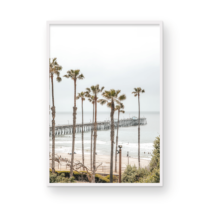 A vertical landscape coastal photography shot of the San Clemente Pier from behind a lush line of tall palm trees. This beachscape print highlights the California coast and is ideal for large beach art or a beach theme wall decor, with subtle ocean waves. Photograph in minimalist mock up.