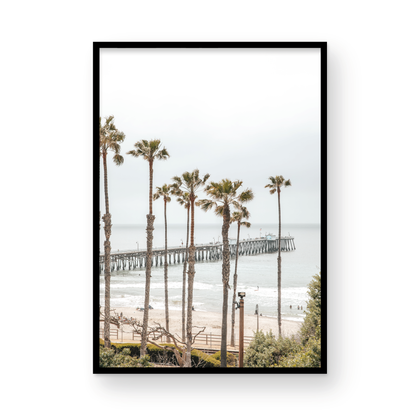 A vertical landscape coastal photography shot of the San Clemente Pier from behind a lush line of tall palm trees. This beachscape print highlights the California coast and is ideal for large beach art or a beach theme wall decor, with subtle ocean waves. Photograph in minimalist mock up.