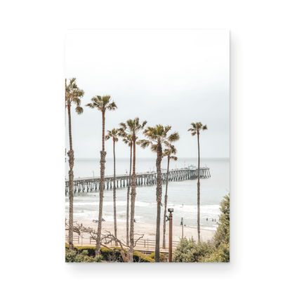 A vertical landscape coastal photography shot of the San Clemente Pier from behind a lush line of tall palm trees. This beachscape print highlights the California coast and is ideal for large beach art or a beach theme wall decor, with subtle ocean waves. Photograph in minimalist mock up.