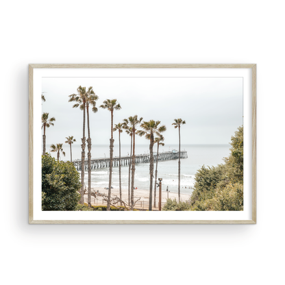A horizontal landscape coastal photography shot of the San Clemente Pier from behind a lush line of tall palm trees. This beachscape print highlights the California coast and is ideal for large landscape beach art or a beach theme wall decor, with subtle ocean waves. Photograph in minimalist mock up.