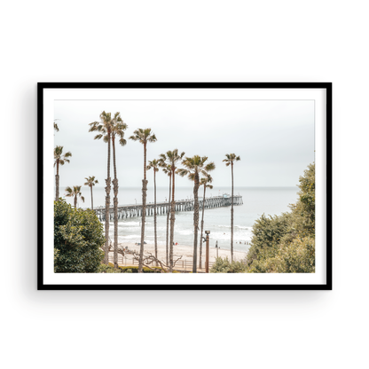 A horizontal landscape coastal photography shot of the San Clemente Pier from behind a lush line of tall palm trees. This beachscape print highlights the California coast and is ideal for large landscape beach art or a beach theme wall decor, with subtle ocean waves. Photograph in minimalist mock up.