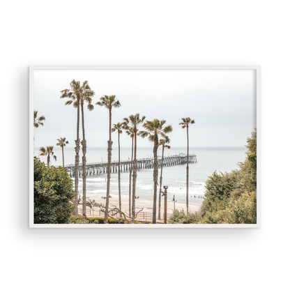 A horizontal landscape coastal photography shot of the San Clemente Pier from behind a lush line of tall palm trees. This beachscape print highlights the California coast and is ideal for large landscape beach art or a beach theme wall decor, with subtle ocean waves. Photograph in minimalist mock up.