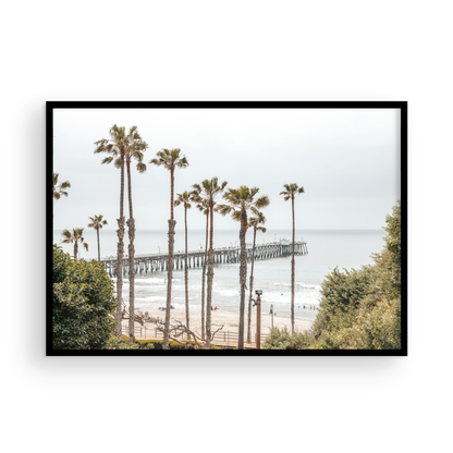 A horizontal landscape coastal photography shot of the San Clemente Pier from behind a lush line of tall palm trees. This beachscape print highlights the California coast and is ideal for large landscape beach art or a beach theme wall decor, with subtle ocean waves. Photograph in minimalist mock up.