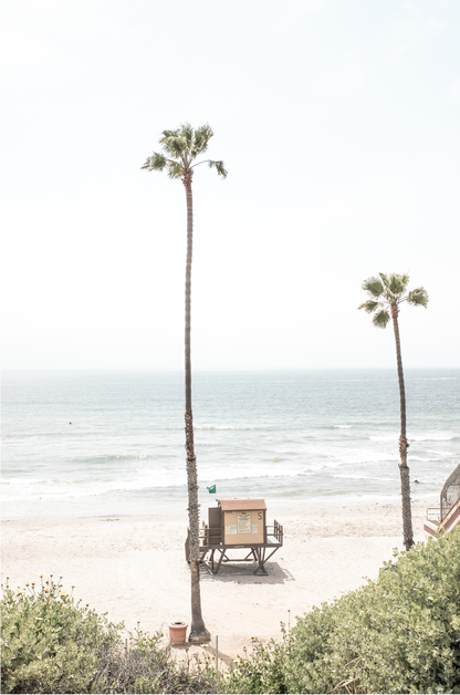 Vertical beach wall art featuring a lifeguard tower nestled between two tall palm trees on a sunny California beach, capturing a serene beach vibe.