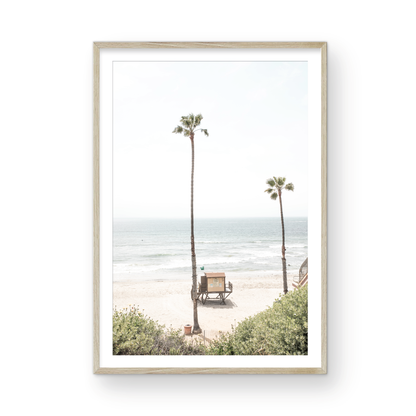 Vertical beach wall art featuring a lifeguard tower nestled between two tall palm trees on a sunny California beach, capturing a serene beach vibe. Photograph in minimalist mock up.