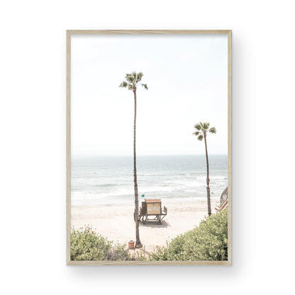 Vertical beach wall art featuring a lifeguard tower nestled between two tall palm trees on a sunny California beach, capturing a serene beach vibe. Photograph in minimalist mock up.