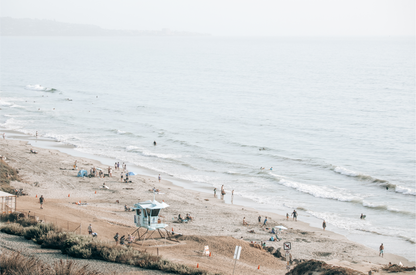 A slightly hazy, high-angle Landscape Beach Print captures a wide beachscape print where a light blue ocean waves meet a sandy shore dotted with numerous people enjoying the Summer Vibes. Gentle white waves roll towards the beach. A Blue Lifeguard Tower stands prominently in the lower center of the framed landscape beach print. The overall lighting suggests a bright, overcast travel photography day on the San Diego coast. 