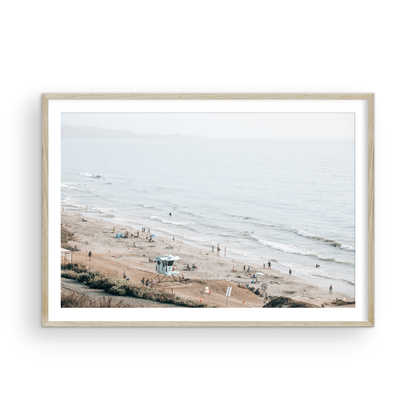 A slightly hazy, high-angle Landscape Beach Print captures a wide beachscape print where a light blue ocean waves meet a sandy shore dotted with numerous people enjoying the Summer Vibes. Gentle white waves roll towards the beach. A Blue Lifeguard Tower stands prominently in the lower center of the framed landscape beach print. The overall lighting suggests a bright, overcast travel photography day on the San Diego coast. Photograph in minimalist mock up