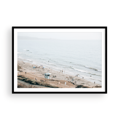 A slightly hazy, high-angle Landscape Beach Print captures a wide beachscape print where a light blue ocean waves meet a sandy shore dotted with numerous people enjoying the Summer Vibes. Gentle white waves roll towards the beach. A Blue Lifeguard Tower stands prominently in the lower center of the framed landscape beach print. The overall lighting suggests a bright, overcast travel photography day on the San Diego coast. Photograph in minimalist mock up