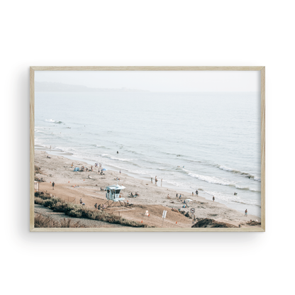 A slightly hazy, high-angle Landscape Beach Print captures a wide beachscape print where a light blue ocean waves meet a sandy shore dotted with numerous people enjoying the Summer Vibes. Gentle white waves roll towards the beach. A Blue Lifeguard Tower stands prominently in the lower center of the framed landscape beach print. The overall lighting suggests a bright, overcast travel photography day on the San Diego coast. Photograph in minimalist mock up