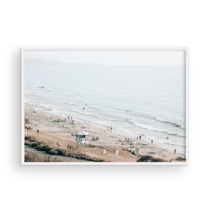 A slightly hazy, high-angle Landscape Beach Print captures a wide beachscape print where a light blue ocean waves meet a sandy shore dotted with numerous people enjoying the Summer Vibes. Gentle white waves roll towards the beach. A Blue Lifeguard Tower stands prominently in the lower center of the framed landscape beach print. The overall lighting suggests a bright, overcast travel photography day on the San Diego coast. Photograph in minimalist mock up