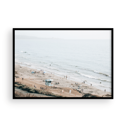 A slightly hazy, high-angle Landscape Beach Print captures a wide beachscape print where a light blue ocean waves meet a sandy shore dotted with numerous people enjoying the Summer Vibes. Gentle white waves roll towards the beach. A Blue Lifeguard Tower stands prominently in the lower center of the framed landscape beach print. The overall lighting suggests a bright, overcast travel photography day on the San Diego coast. Photograph in minimalist mock up