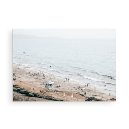 A slightly hazy, high-angle Landscape Beach Print captures a wide beachscape print where a light blue ocean waves meet a sandy shore dotted with numerous people enjoying the Summer Vibes. Gentle white waves roll towards the beach. A Blue Lifeguard Tower stands prominently in the lower center of the framed landscape beach print. The overall lighting suggests a bright, overcast travel photography day on the San Diego coast. Photograph in minimalist mock up