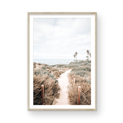Minimalist vertical beach print showcasing the San Diego Sunset Cliffs, a sandy pathway with rustic wooden steps leading through coastal dune grass towards a calm ocean. This pastel beach art, evocative of the Southern California coastline, features a bright hazy sky and distant palm trees. A tranquil piece of travel photography, ideal for coastal wall art. Photograph in minimalist framed mock up.