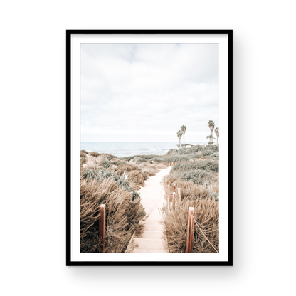 Minimalist vertical beach print showcasing the San Diego Sunset Cliffs, a sandy pathway with rustic wooden steps leading through coastal dune grass towards a calm ocean. This pastel beach art, evocative of the Southern California coastline, features a bright hazy sky and distant palm trees. A tranquil piece of travel photography, ideal for coastal wall art. Photograph in minimalist framed mock up.