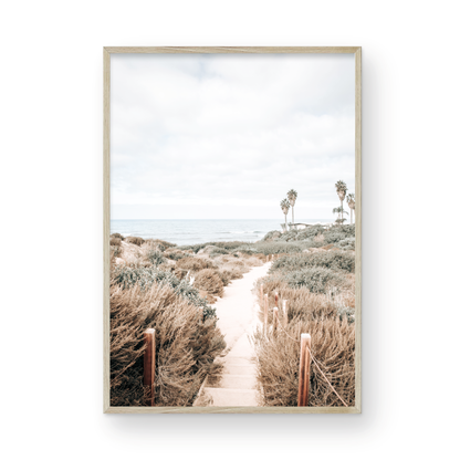 Minimalist vertical beach print showcasing the San Diego Sunset Cliffs, a sandy pathway with rustic wooden steps leading through coastal dune grass towards a calm ocean. This pastel beach art, evocative of the Southern California coastline, features a bright hazy sky and distant palm trees. A tranquil piece of travel photography, ideal for coastal wall art. Photograph in minimalist framed mock up.