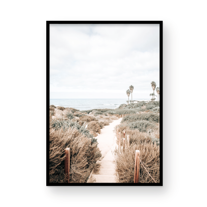 Minimalist vertical beach print showcasing the San Diego Sunset Cliffs, a sandy pathway with rustic wooden steps leading through coastal dune grass towards a calm ocean. This pastel beach art, evocative of the Southern California coastline, features a bright hazy sky and distant palm trees. A tranquil piece of travel photography, ideal for coastal wall art. Photograph in minimalist framed mock up.