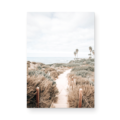 Minimalist vertical beach print showcasing the San Diego Sunset Cliffs, a sandy pathway with rustic wooden steps leading through coastal dune grass towards a calm ocean. This pastel beach art, evocative of the Southern California coastline, features a bright hazy sky and distant palm trees. A tranquil piece of travel photography, ideal for coastal wall art. Photograph in minimalist unframed mock up.