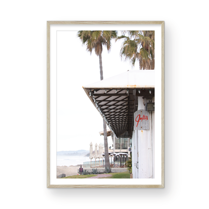 A framed beach scene art print with a wooden structure and palm trees, titled 'Jake's' on the sign, set against a background of a beach and ocean. Photograph in minimalist mock up.