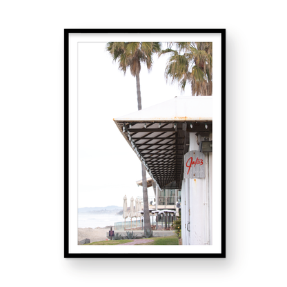 A framed beach scene art print with a wooden structure and palm trees, titled 'Jake's' on the sign, set against a background of a beach and ocean. Photograph in minimalist mock up.