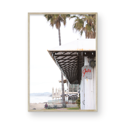 A framed beach scene art print with a wooden structure and palm trees, titled 'Jake's' on the sign, set against a background of a beach and ocean. Photograph in minimalist mock up.