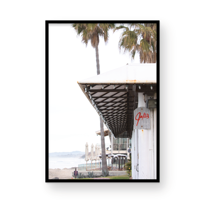 A framed beach scene art print with a wooden structure and palm trees, titled 'Jake's' on the sign, set against a background of a beach and ocean. Photograph in minimalist mock up.