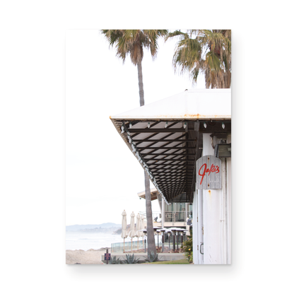 A framed beach scene art print with a wooden structure and palm trees, titled 'Jake's' on the sign, set against a background of a beach and ocean. Photograph in minimalist mock up.