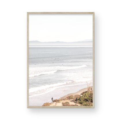 Minimalist ocean print of ocean waves with surfers in the distance and a sandy hillside in the foreground. 