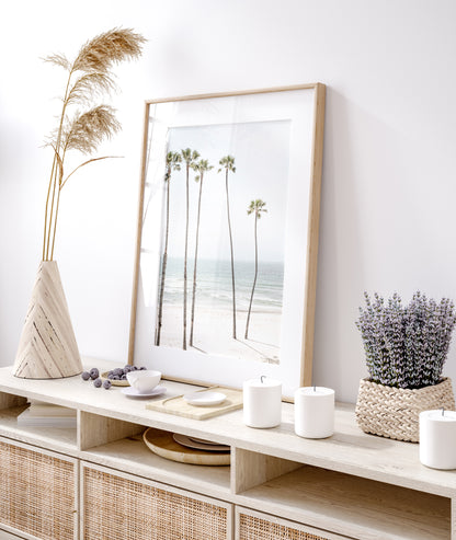 Vertical coastal print showcasing a line of very tall palm trees against the bright sky, overlooking the calm ocean waves. A quintessential tropical coastal print. Photograph in minimalist mock up.