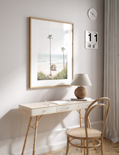 Vertical beach wall art featuring a lifeguard tower nestled between two tall palm trees on a sunny California beach, capturing a serene beach vibe. Photograph in minimalist mock up.