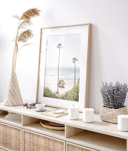 Vertical beach wall art featuring a lifeguard tower nestled between two tall palm trees on a sunny California beach, capturing a serene beach vibe. Photograph in minimalist mock up.