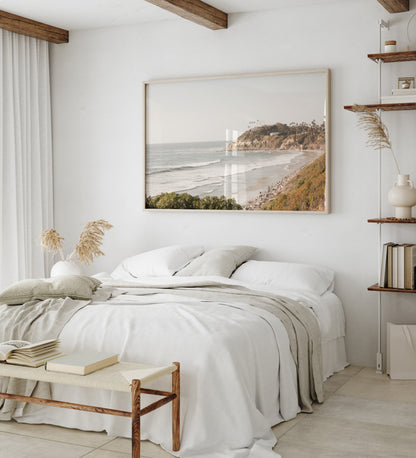 Fine art of Swamis Beach, San Diego California horizontal print of hazy high-angle landscape beach photography captures a light blue ocean meeting a sandy beach with many beach goers. To the right, tall tan and brown cliffs rise as iconic coastal scenery. Photograph in minimalist framed mock up.
