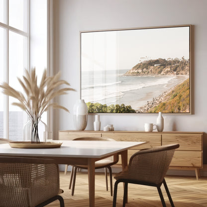 Fine art of Swamis Beach, San Diego California horizontal print of hazy high-angle landscape beach photography captures a light blue ocean meeting a sandy beach with many beach goers. To the right, tall tan and brown cliffs rise as iconic coastal scenery. Photograph in minimalist framed mock up.
