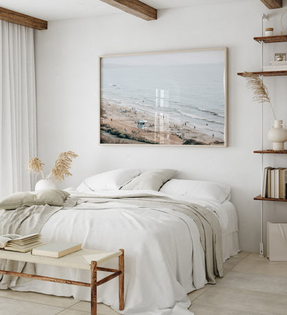 A slightly hazy, high-angle Landscape Beach Print captures a wide beachscape print where a light blue ocean waves meet a sandy shore dotted with numerous people enjoying the Summer Vibes. Gentle white waves roll towards the beach. A Blue Lifeguard Tower stands prominently in the lower center of the framed landscape beach print. The overall lighting suggests a bright, overcast travel photography day on the San Diego coast. Photograph in minimalist mock up